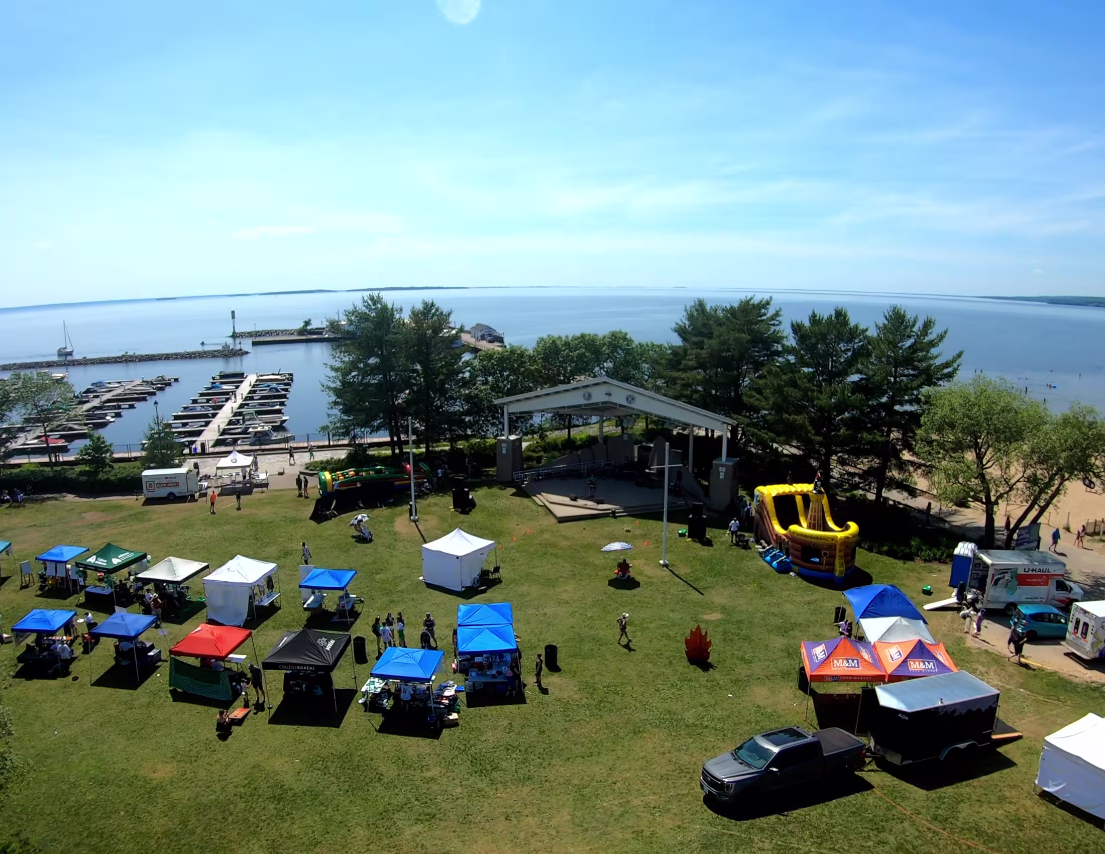 Photo drone montrant le Kiwanis Bandshell et le parc devant. Il y a une variété de tentes d'événements et à l'horizon, on voit le lac Nipissing.