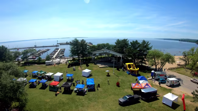 Photo drone montrant le Kiwanis Bandshell et le parc devant. Il y a une variété de tentes d'événements et à l'horizon, on voit le lac Nipissing.